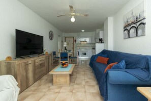 Sala de estar com televisão, móveis de madeira, sofá azul, ventilador de teto e cozinha ao fundo.