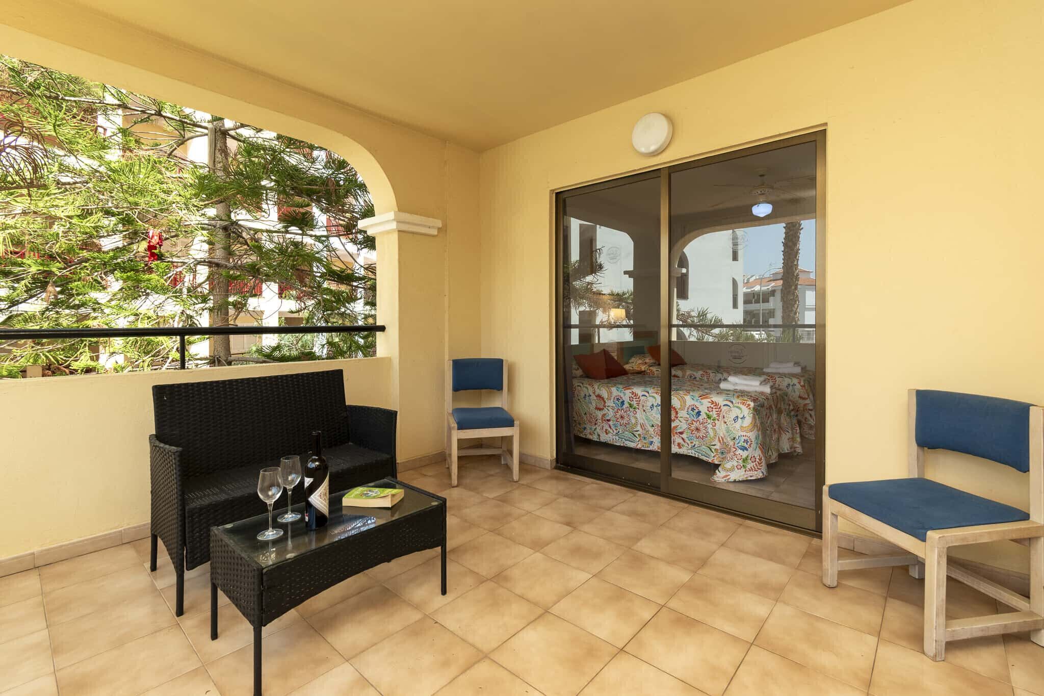 Un balcon aménagé avec un canapé, des chaises, une table et une porte-fenêtre coulissante donnant sur une chambre à coucher équipée d’un lit et d’une table.