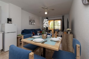 A dining area with a table set for two, a refrigerator, a sofa, and a view of the outdoors.