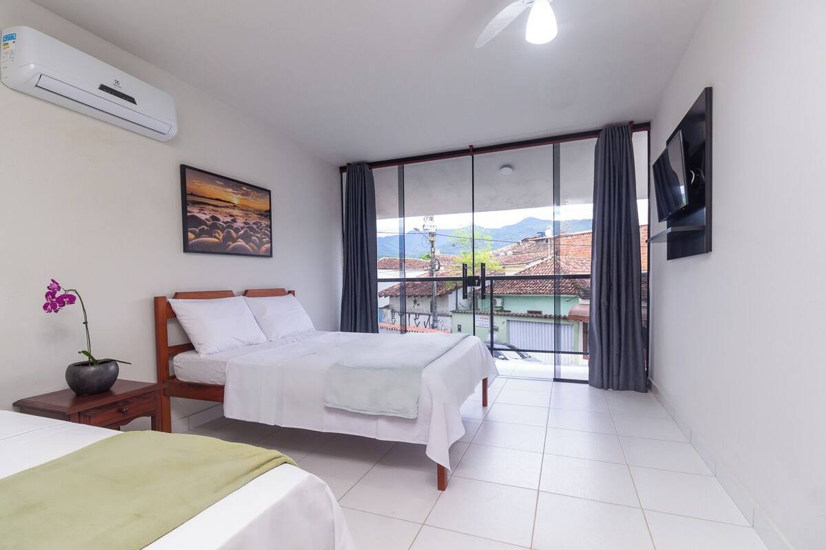 A hotel room with a bed, a small table with a plant, a painting, a TV, and a balcony with a view.