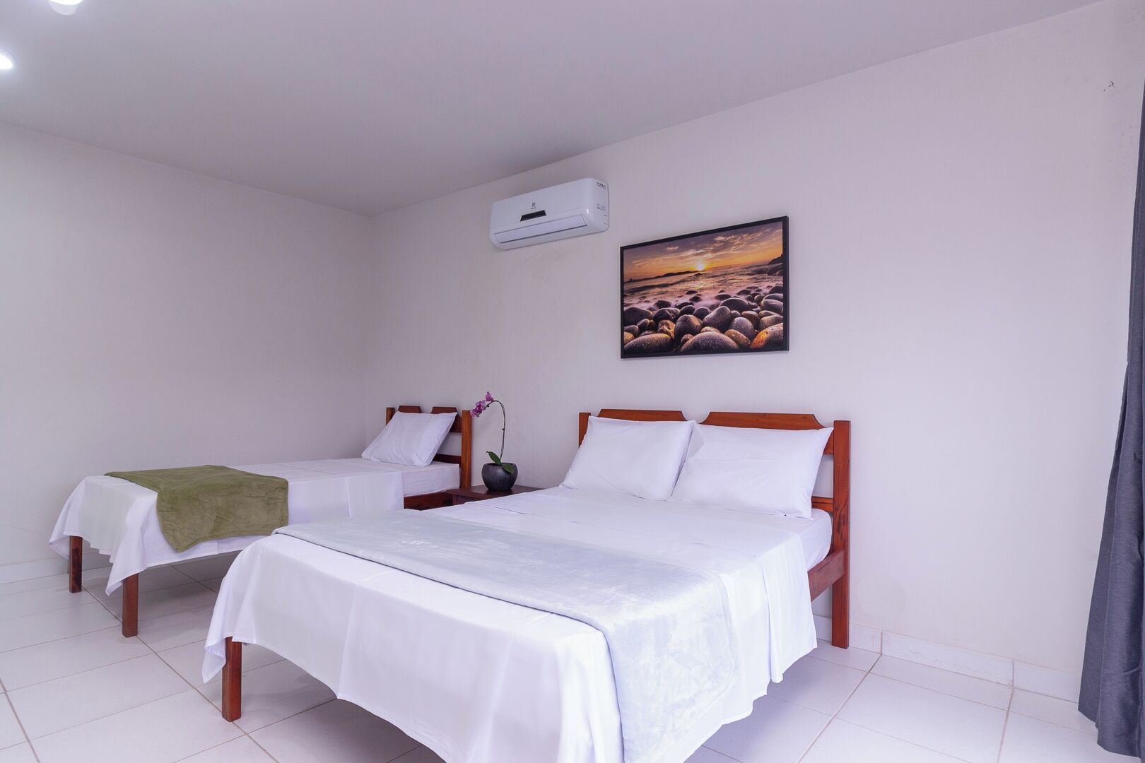 A hotel room with two single beds, a wall-mounted air conditioner, and a painting of a sunset over rocks.