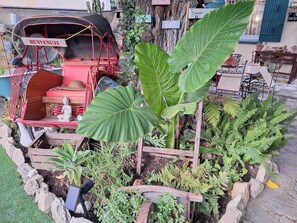 Ein Gartenbereich mit einem roten Wagen, der „BENVENUTI“ zeigt, umgeben von verschiedenen Pflanzen und einer kleinen Statue.