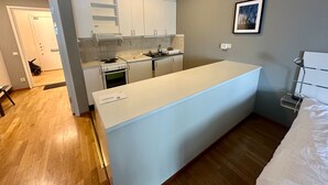 Une cuisine compacte avec des meubles blancs, une cuisinière et un évier. Un tableau encadré est accroché au mur et un petit banc se trouve dans le coin.