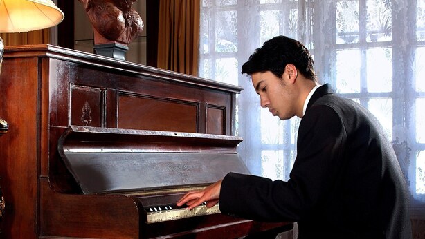 A person playing a grand piano in a room with a large window and a decorative bust on the wall. - Welcomheritage Kasmanda Palace (Dehradun)