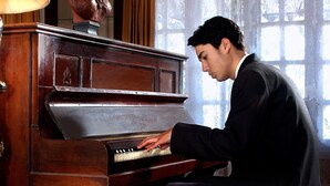 Una persona suona un pianoforte a coda in una stanza con un'ampia finestra e un busto decorativo appeso al muro.