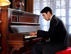 A person playing a grand piano in a room with a large window and a decorative bust on the wall.