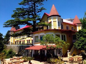 A large, multi-story building with a distinctive red roof and yellow walls, featuring a balcony with outdoor furniture. - Welcomheritage Kasmanda Palace (Dehradun)