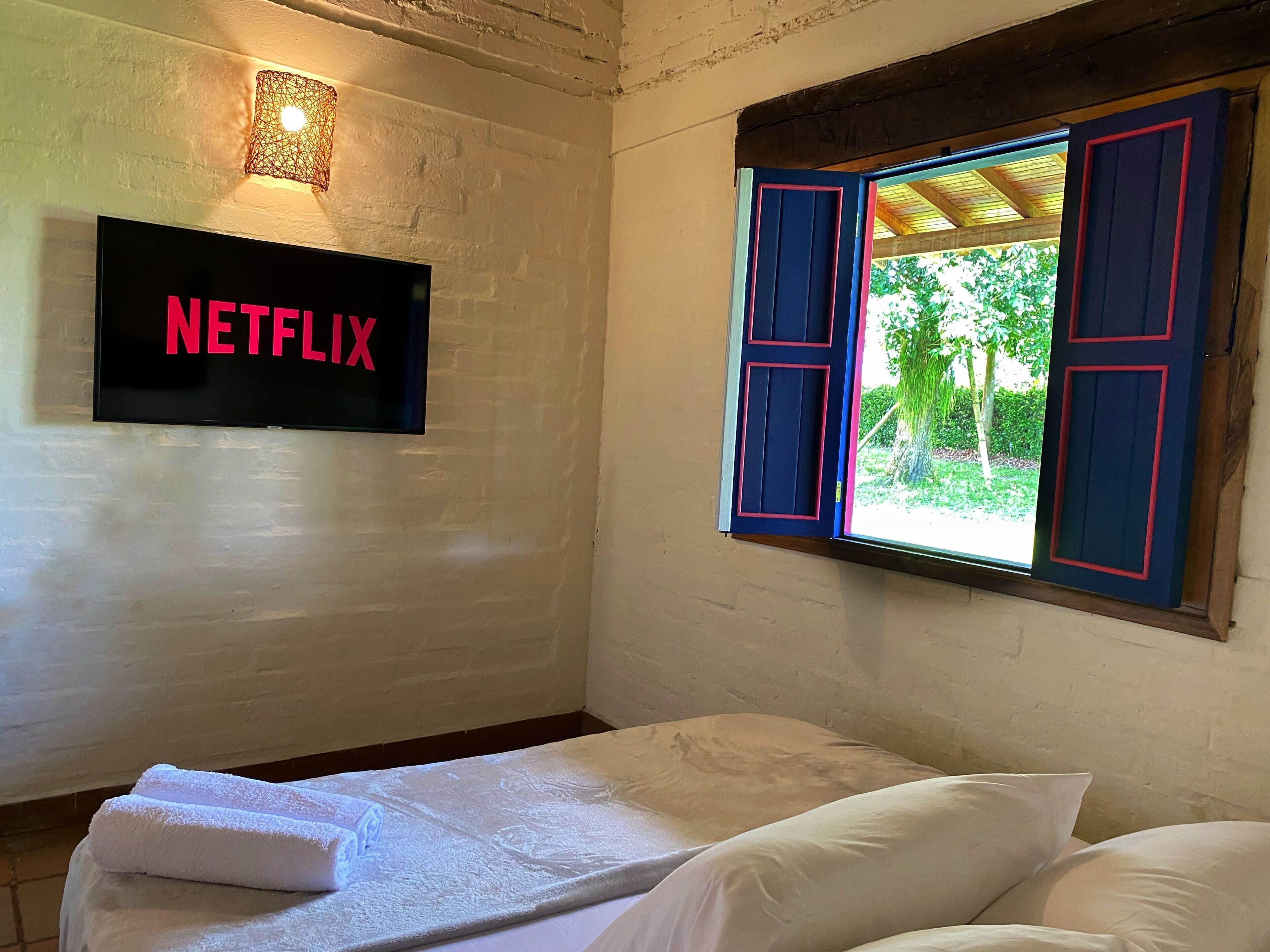 Una habitación con cama, un televisor de pared mostrando 'NETFLIX' y una ventana con contraventanas azules.