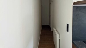 A narrow hallway with white walls and a wooden floor. A radiator is mounted on the wall, and a door is visible at the end of the hallway.