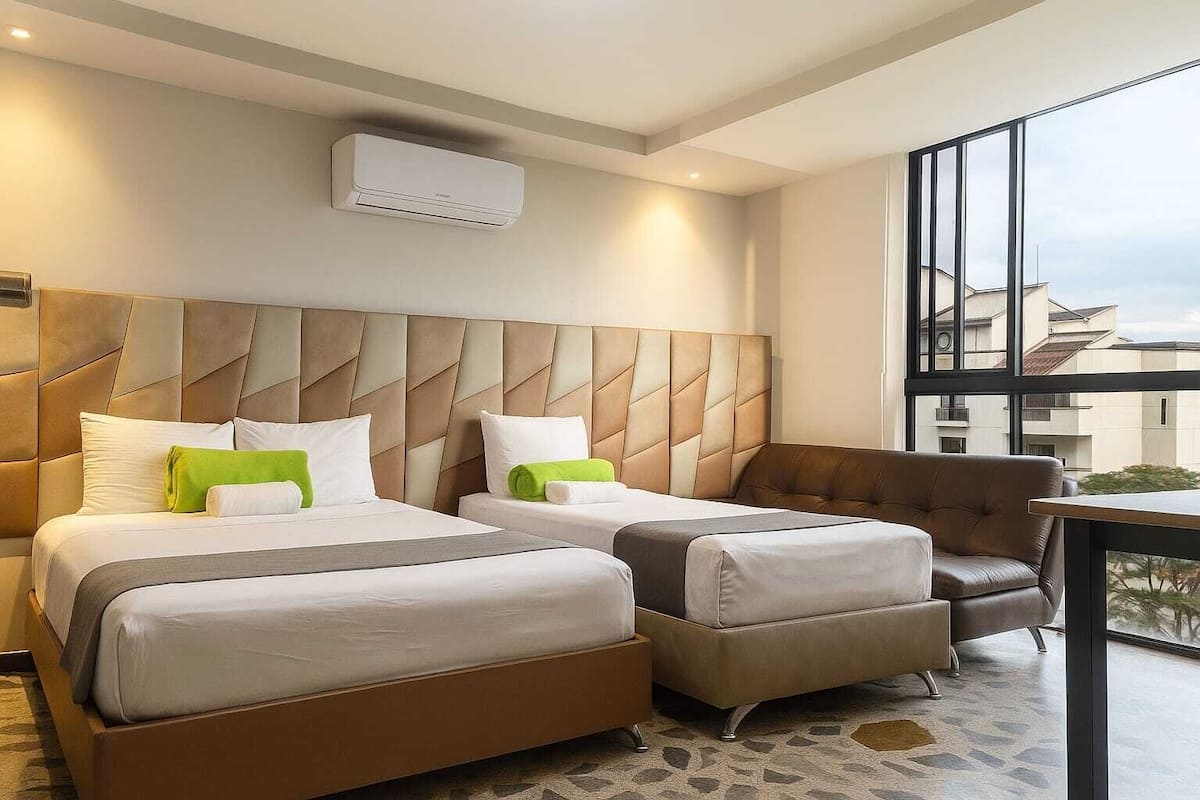 A hotel room with two beds, a sofa, a small table, and a large window offering a view of buildings.