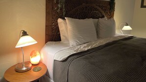Un lit bien fait, avec du linge de lit blanc, une couverture à motifs et une tête de lit décorative ornée d’un motif floral. On y voit deux lampes de chevet, l’une avec un pied en forme de globe et l’autre avec un pied plat. Une petite table ronde, sur laquelle se trouvent un globe et une lampe, est visible à gauche du lit.