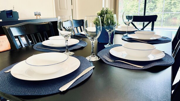Une table à manger dressée pour quatre personnes, avec des sets de table bleus, des assiettes blanches et de la verrerie transparente.