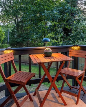 Eine Holzterrasse mit Tisch und zwei Stühlen, umgeben von einem Geländer und viel Grün.