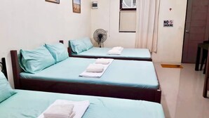 A hotel room with two beds, each with a light blue mattress and white pillows. There is a fan on the wall, a "NO SMOKING" sign, and a door with a cross symbol.
