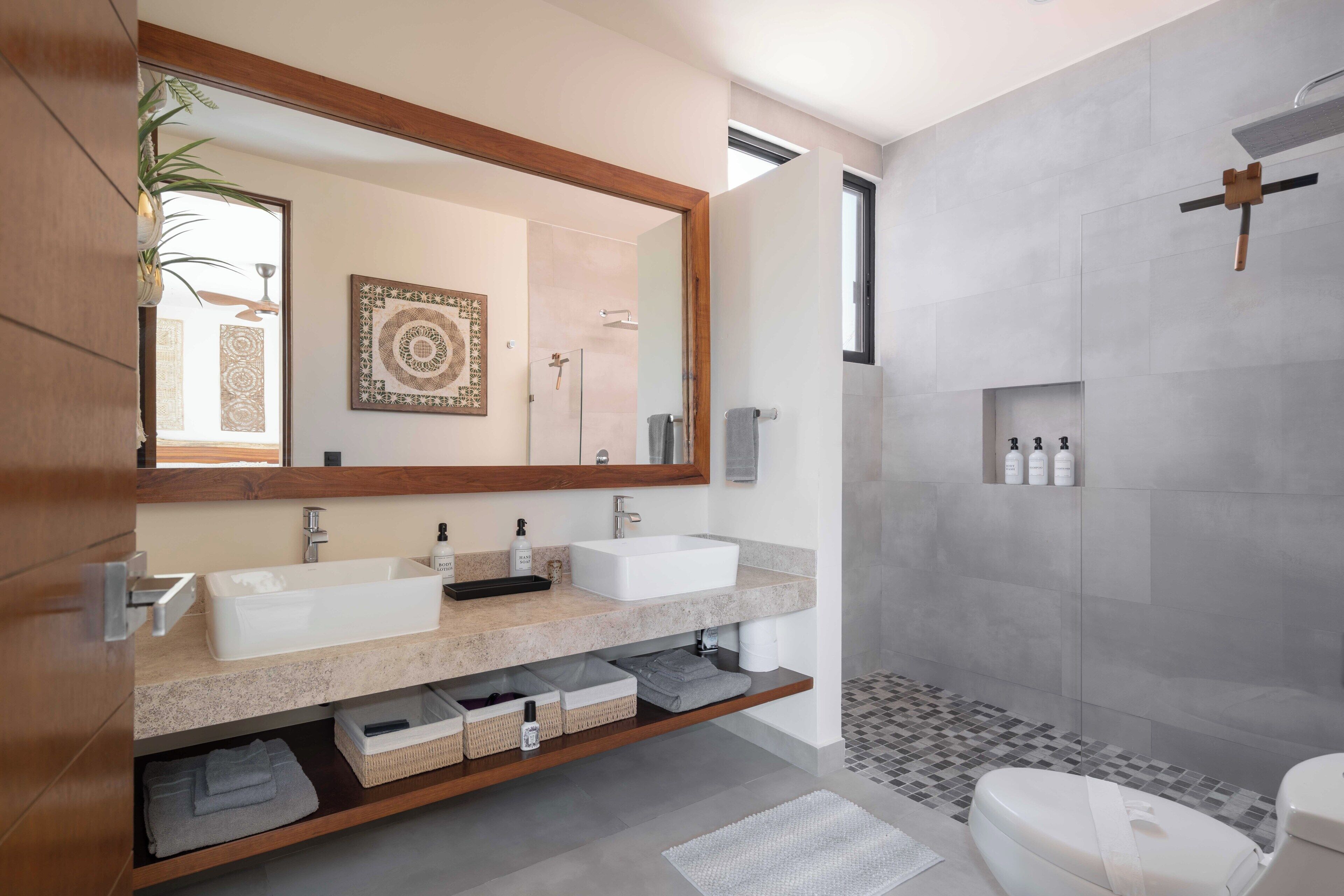 Une salle de bain moderne avec un grand miroir, deux lavabos, une douche et des toilettes. On trouve des paniers de rangement sous les lavabos et une petite étagère avec des articles de toilette.