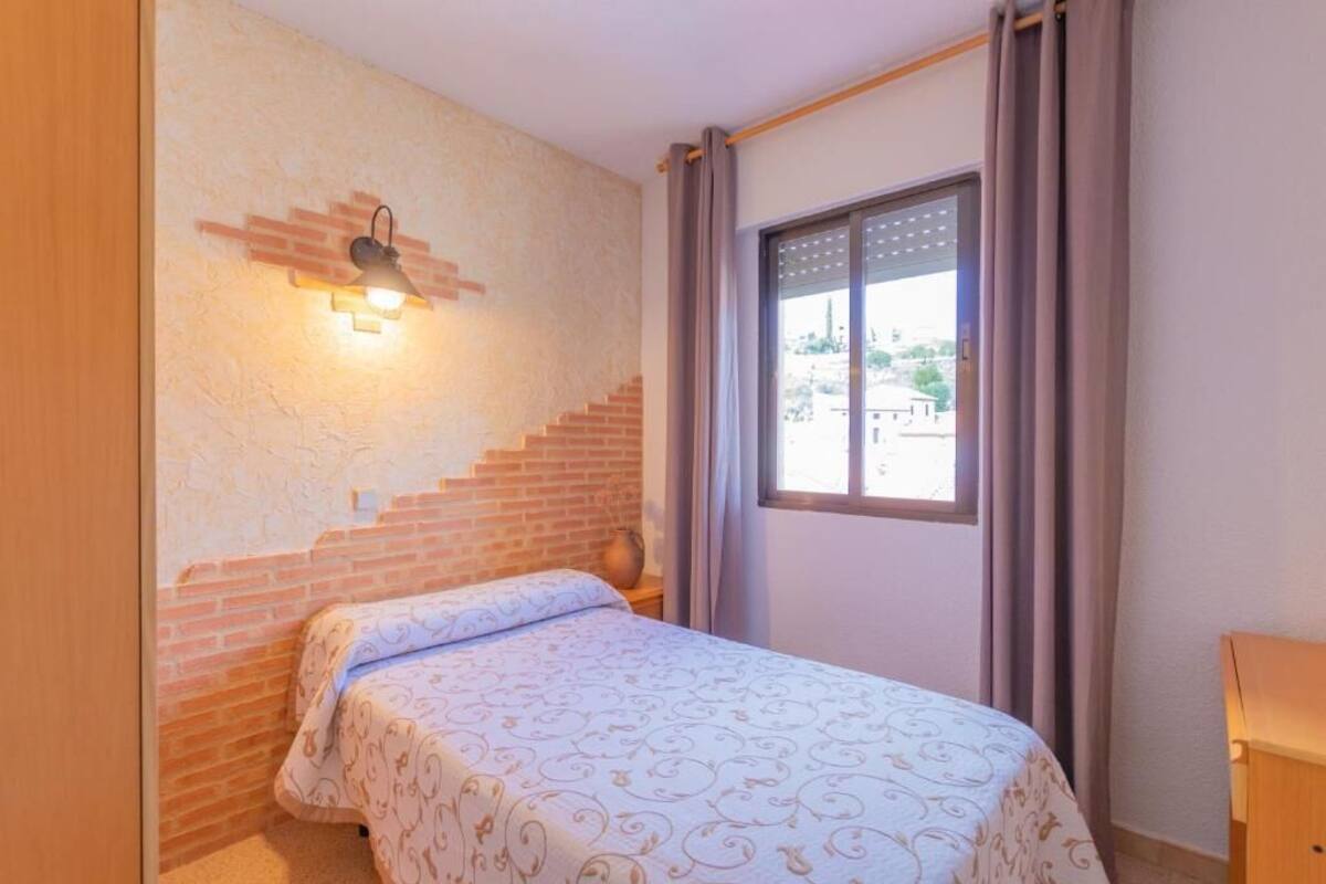 A bedroom with a bed, a window with curtains, a brick wall, and a wall-mounted light fixture.