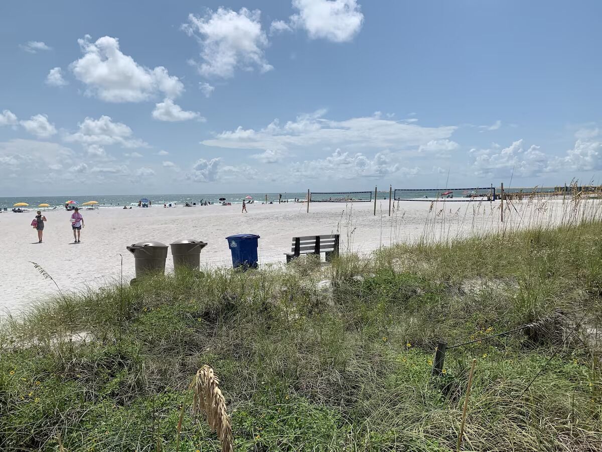 Een strand met zand, gras en enkele personen. Vuilnisbakken en een bankje zijn aanwezig.