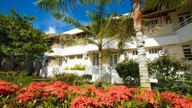 A white building with a thatched roof, surrounded by lush greenery and red flowers. - Pousada Aruana Praia (Aracaju)