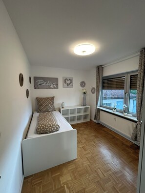 Un dormitorio con cama, una consola blanca, adornos en la pared, una ventana con cortinas y un radiador.