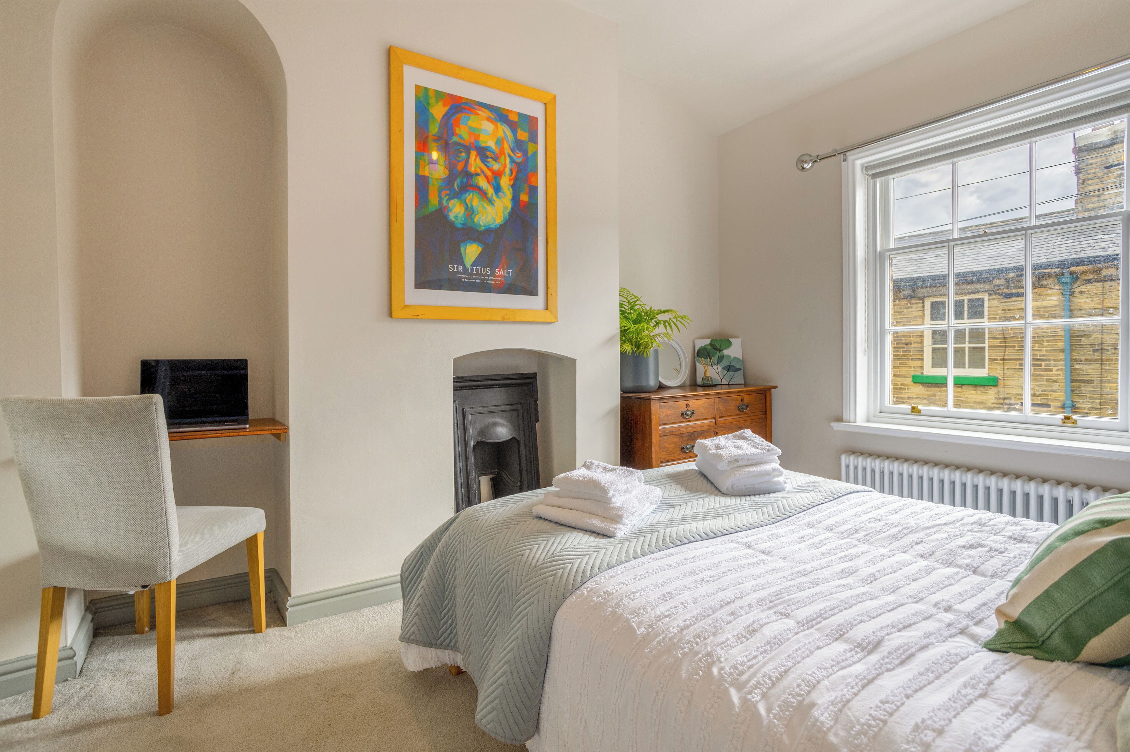 A bedroom with a bed, a chair, a fireplace, a window, and a painting on the wall.