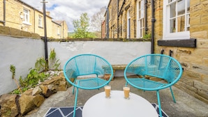 A small outdoor seating area with two turquoise chairs and a white table, set against a stone wall with a window and a brick building in the background. - Stylish Saltaire Cottage | Walk to Salts Mill | Parking & Fast WiFi (Saltaire)