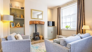 A cozy living room with two grey sofas, a fireplace, a TV, and a large window with curtains. - Stylish Saltaire Cottage | Walk to Salts Mill | Parking & Fast WiFi (Saltaire)