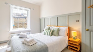 A bedroom with a large window, a bed with white bedding, a bedside lamp, and a radiator. - Stylish Saltaire Cottage | Walk to Salts Mill | Parking & Fast WiFi (Saltaire)
