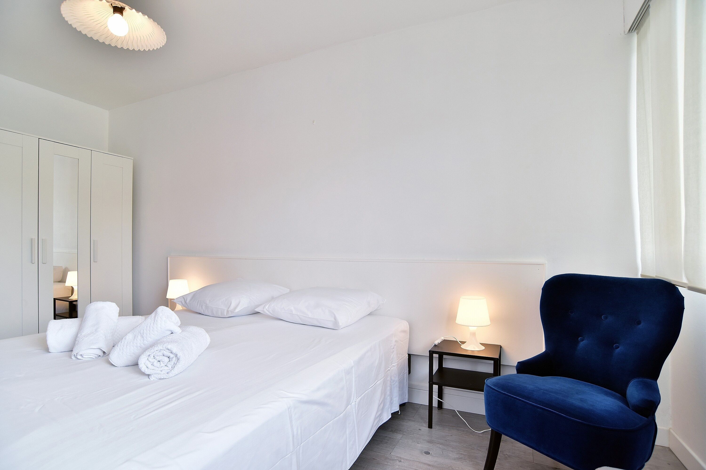 A hotel room with a neatly made bed, a blue armchair, a bedside table with a lamp, and a wardrobe.