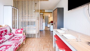 Une chambre d’hôtel moderne avec un plancher en bois, un canapé agrémenté de coussins à motifs, une table à manger dressée avec des assiettes et des verres, et un téléviseur fixé au mur.