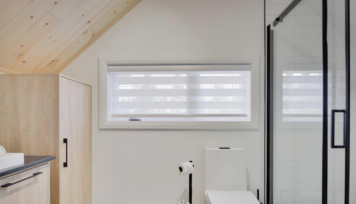 A modern bathroom with a shower, a toilet, and a window with blinds.
