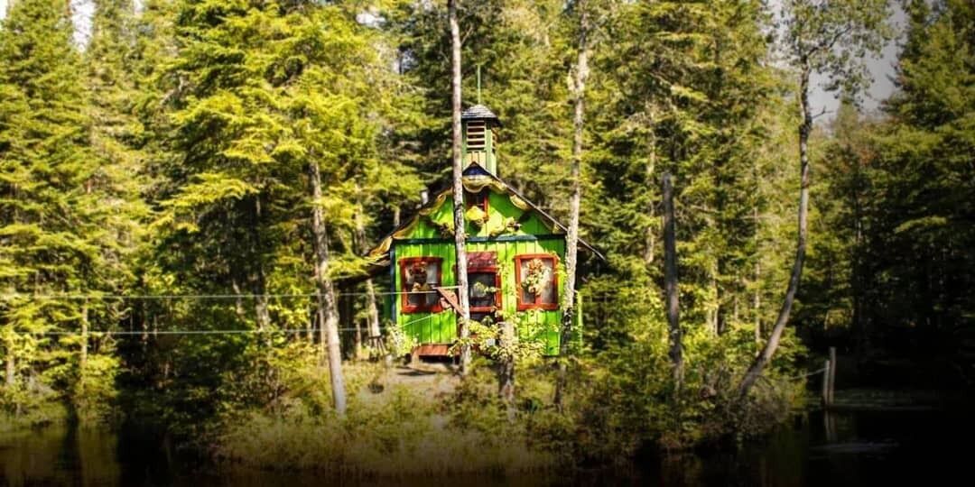 Une construction ressemblant à un chalet, entourée d’une forêt dense, avec un petit quai s’avançant dans une étendue d’eau.
