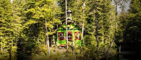 Une construction ressemblant à un chalet, entourée d’une forêt dense, avec un petit quai s’avançant dans une étendue d’eau.