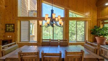 A wooden interior with a large dining table and benches, a chandelier, and large windows offering a view of the outdoors.