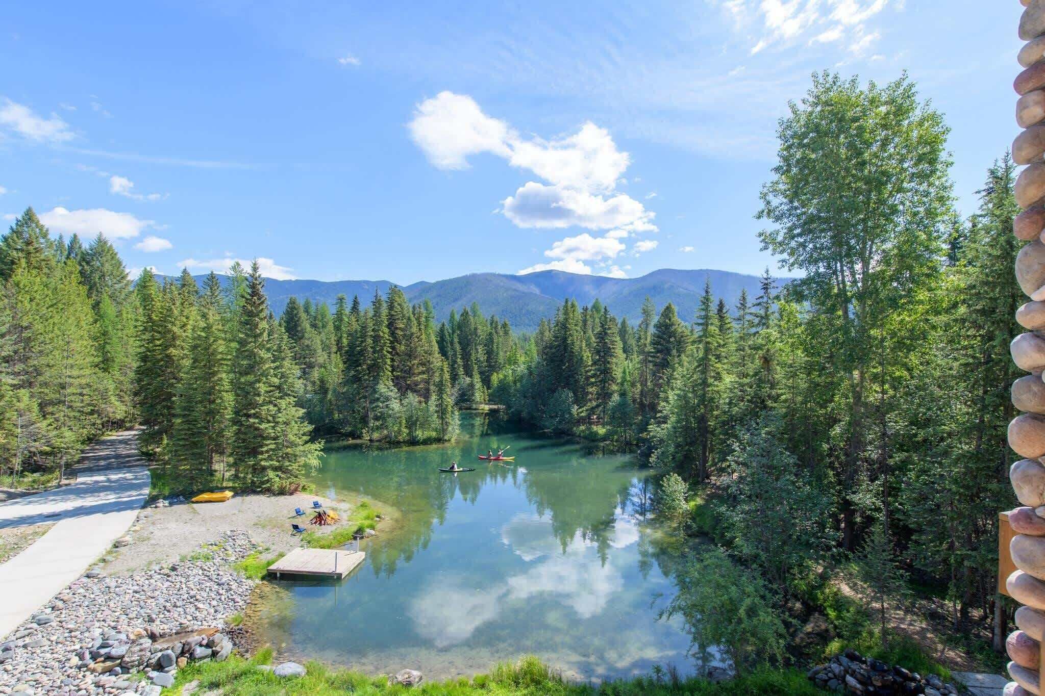 Une vue paisible sur un lac, avec un sentier dégagé menant à un quai, le tout entouré d’une forêt dense et de montagnes en arrière-plan.