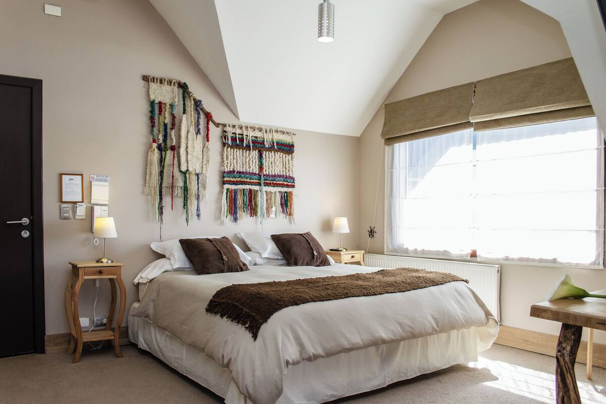 Une chambre avec un grand lit, une table de chevet en bois, une décoration murale en macramé et une fenêtre donnant sur un beau paysage.
