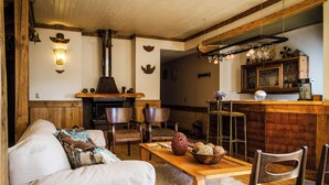 Un salon chaleureux avec un canapé, une table basse en bois et des fauteuils. On aperçoit une cheminée au fond, et des objets décoratifs ornent les murs.