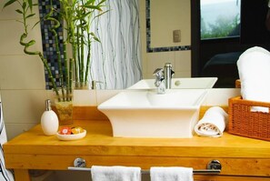 Un baño con lavamanos blanco, un espejo, una planta de bambú, una toalla blanca y una cesta de mimbre.