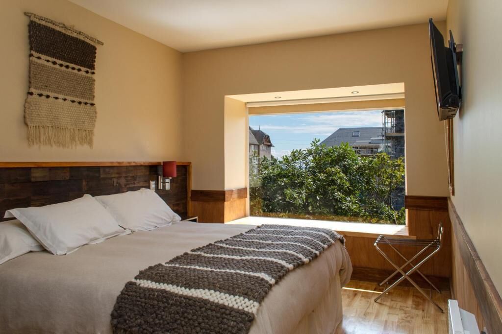 A bedroom with a large bed, a window with a view of greenery and buildings, a flat-screen TV mounted on the wall, and a wooden headboard.