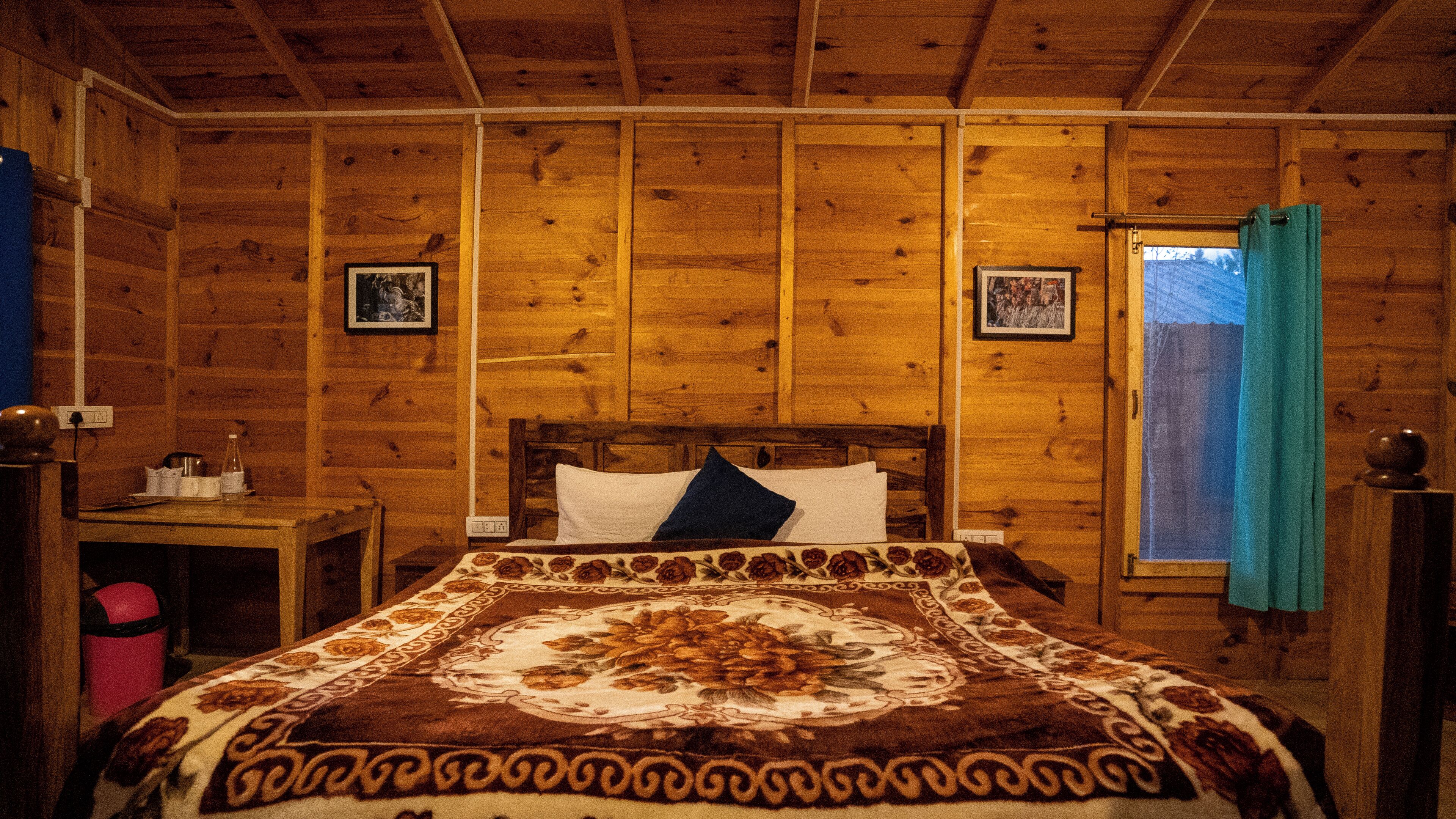 Un dormitorio de cabaña de madera con una cama, una mesita de noche, una ventana con cortinas y dos cuadros en las paredes.