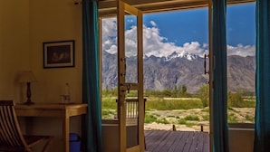 A room with a wooden desk, a chair, a framed picture on the wall, and a door leading to a balcony with a mountain view. - Nubra Ecolodge (Leh)