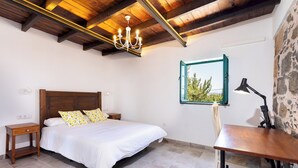A bedroom with a wooden ceiling, a bed with white bedding and patterned pillows, a nightstand, a desk with a lamp, and a stone wall. - Home2Book Rural House with Private Pool & BBQ (El Rosario)
