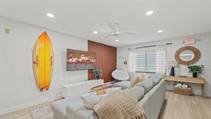 A modern living room with a sofa, a television, a ceiling fan, and a surfboard decoration. - Walk to Beach, 2 Master Beds, Newly Renovated, Walk to Restaurants, Sleeps 8 (Cocoa Beach)