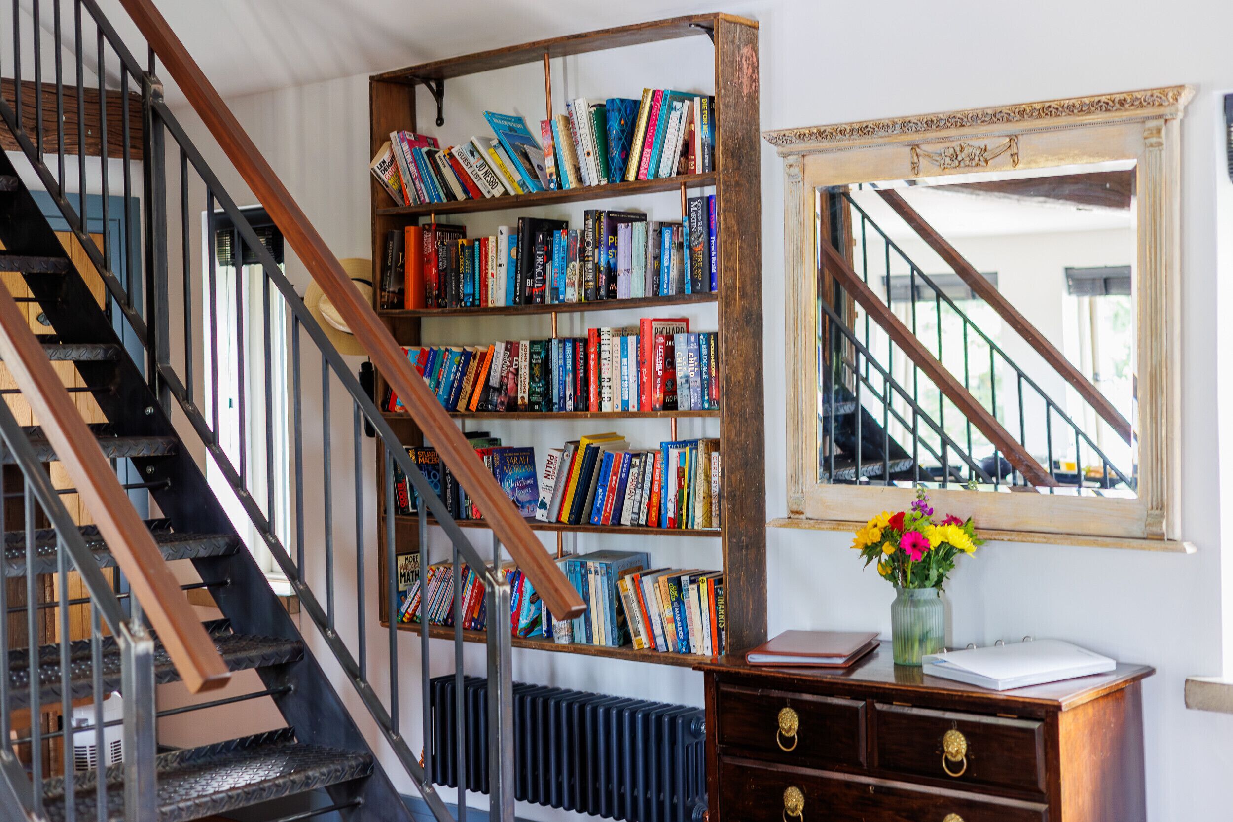 Un escalier avec une rampe en bois, une bibliothèque remplie de livres, un radiateur et un vase de fleurs posé sur une commode en bois.