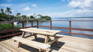 A wooden deck with a picnic table overlooking a calm body of water, surrounded by lush greenery and palm trees. - Wonderful townhouse located directly between the beach and the bay.  Watch the sunsets off the balco (BRADENTON BEACH)