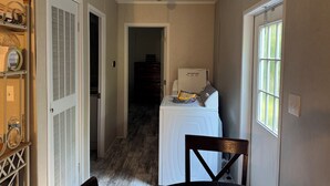 A narrow hallway with a wooden floor, a white door on the left, and a window on the right. There is a small table with a chair in the center.
