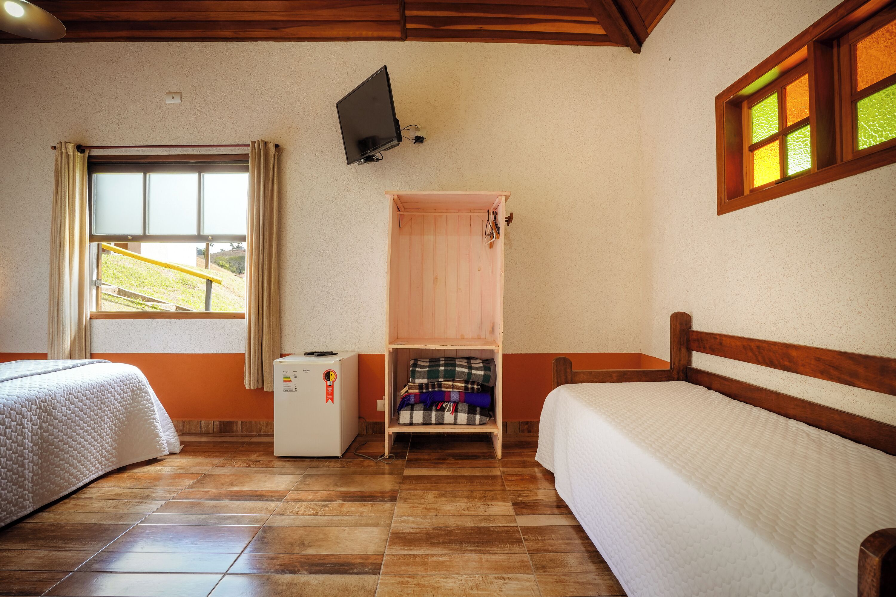 A hotel room with two beds, a TV mounted on the wall, a small desk with a refrigerator, and a window with curtains.