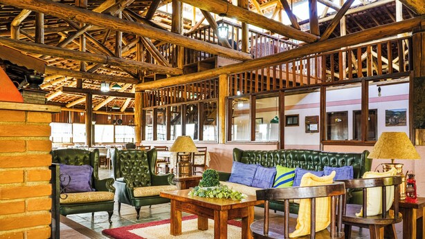 A rustic interior with a high wooden ceiling, leather sofas, wooden tables, and a red carpet.