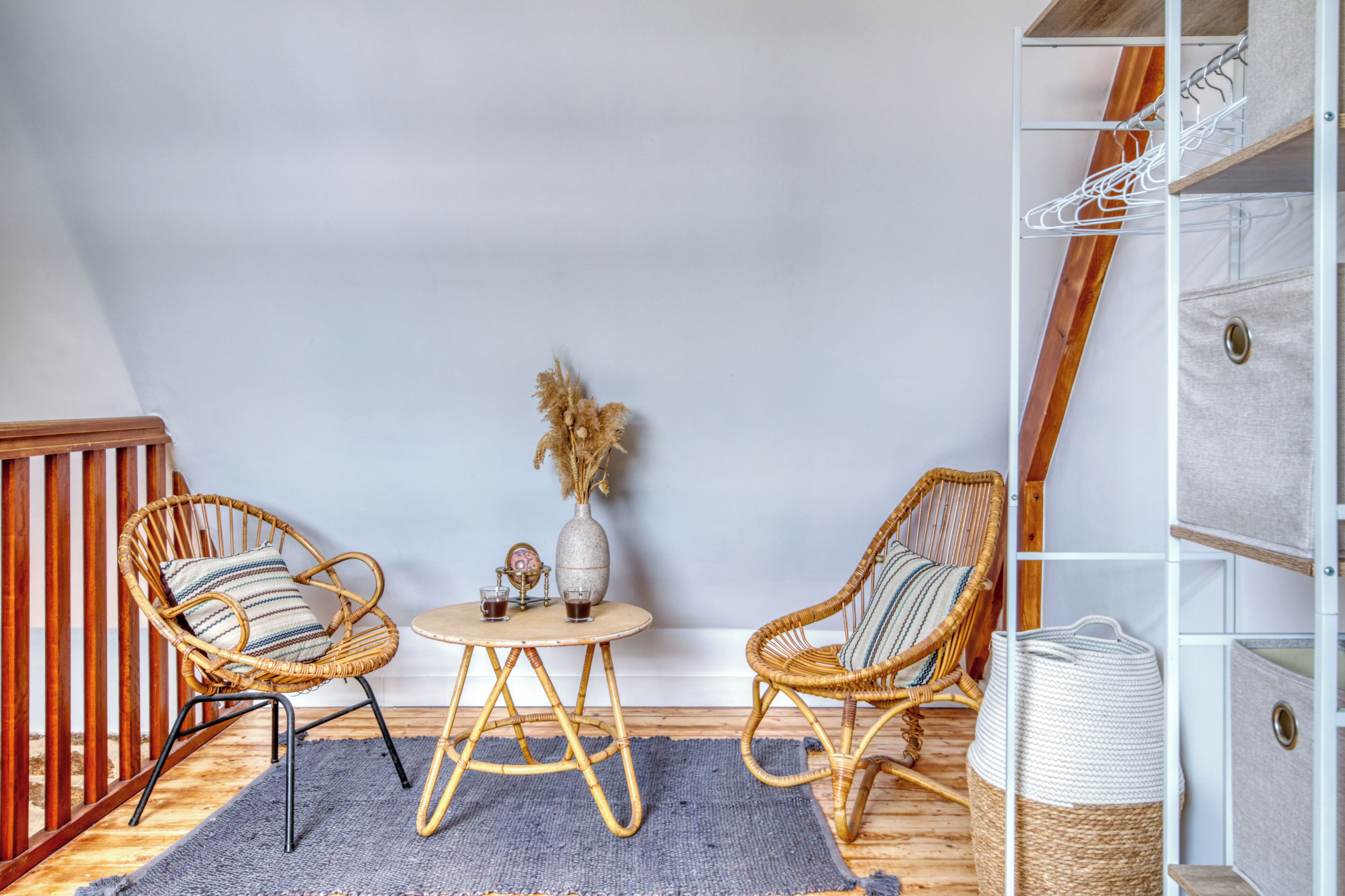 A room with two wicker chairs, a small round table, a vase with dried plants, and a white shelving unit.