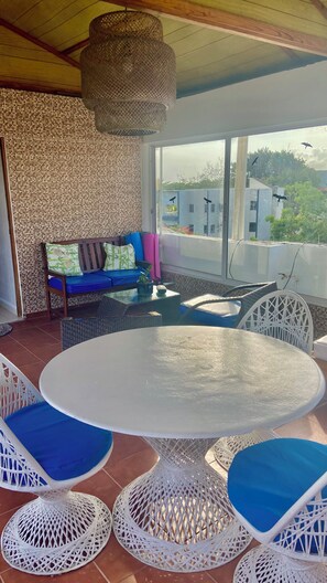 Een overdekt terras met een ronde tafel en witte wickerstoelen, een blauwe bank met kussens en een stenen muur.