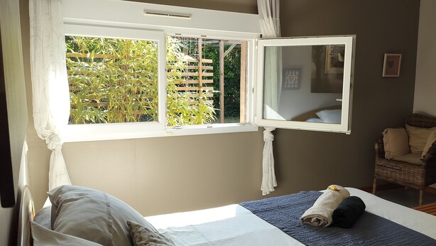 Un dormitorio con cama, ventana con vista a vegetación, una silla de mimbre y un peluche.